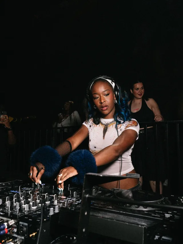 A DJ concentrating on the mix with dancers behind her.